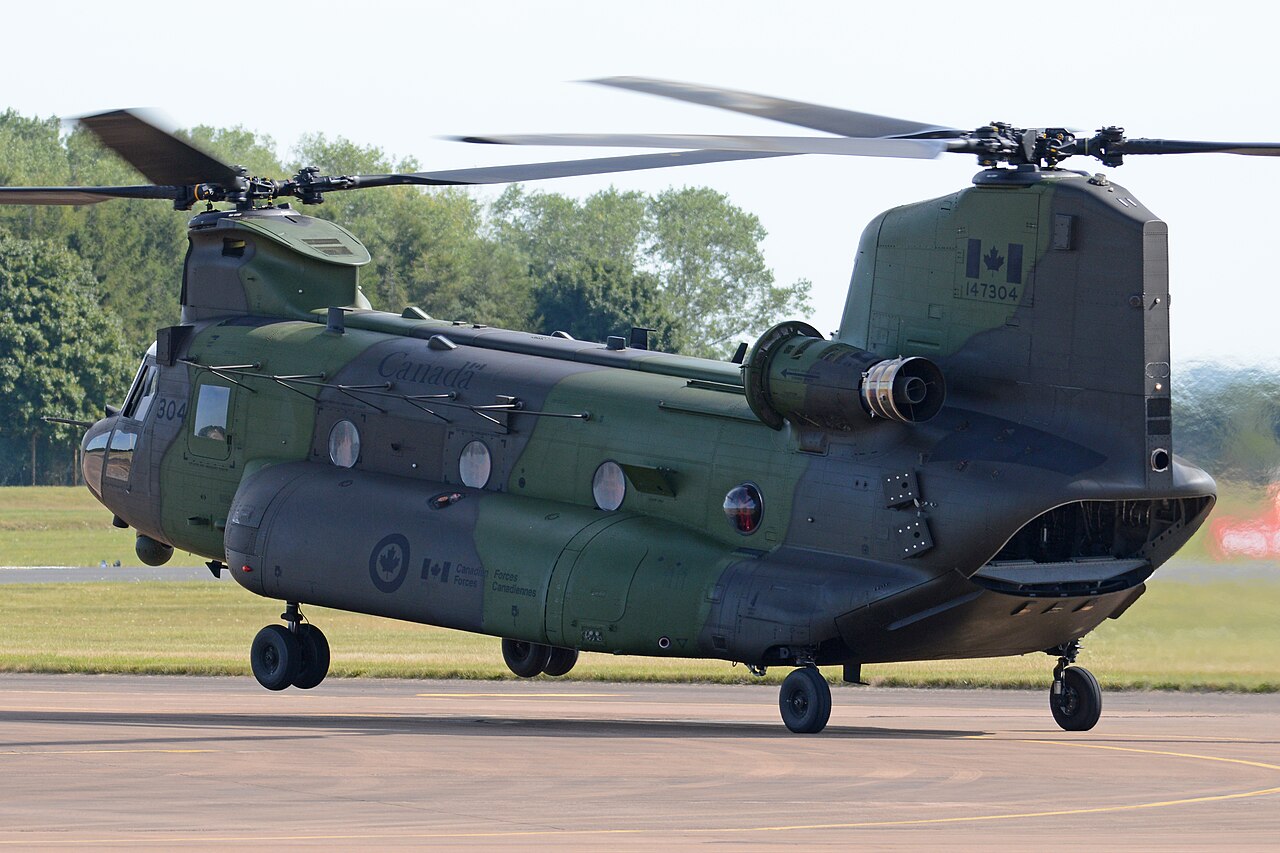 A Canadian CH-147F Chinook with tandem rotors and landing gear extended above an airfield.